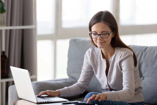 Smiling female math student sit on couch studying on laptop doing research calculating,  positive young woman pay bills online from home make calculations on calculator, check home expenses