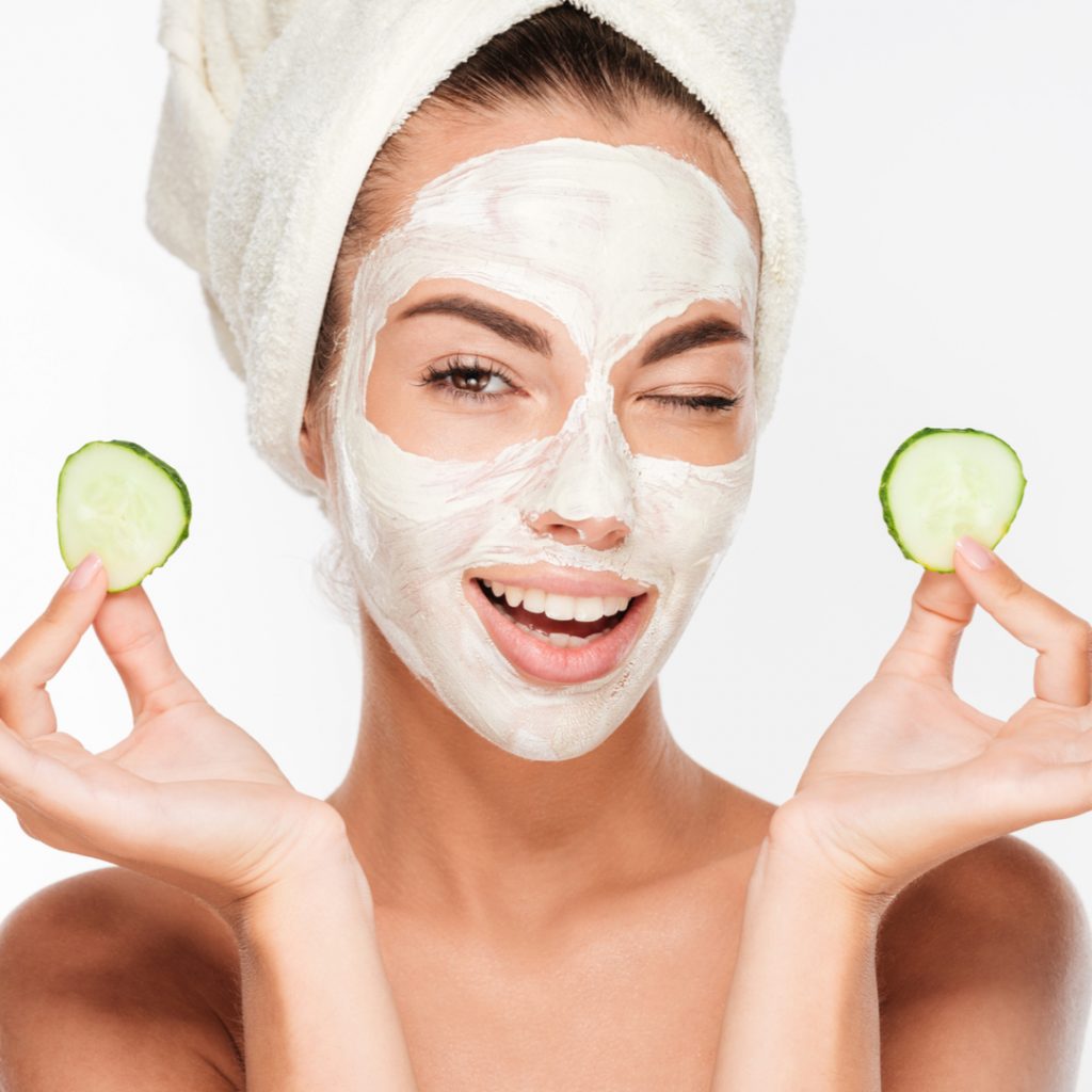 A happy woman wears a beauty mask and places cucumber slices over her eyes.