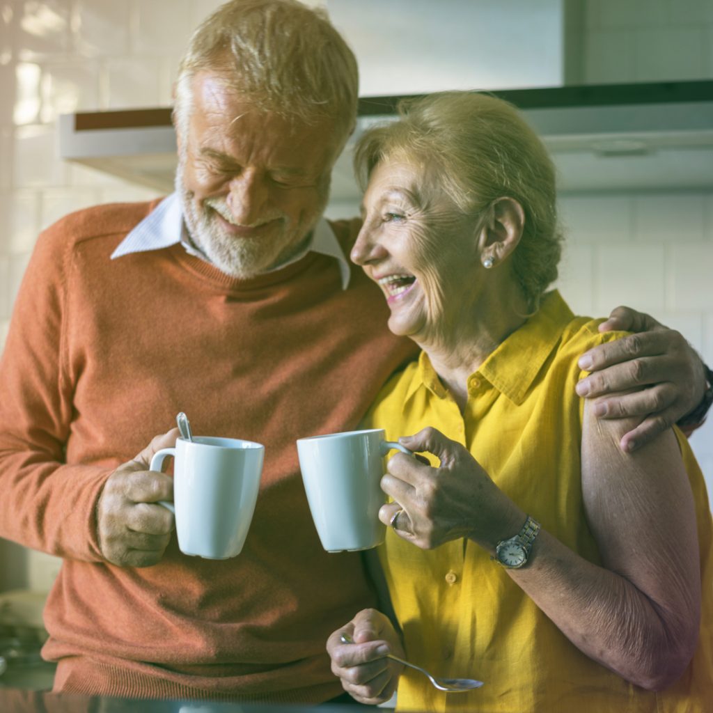 A retired couple enjoys a hug and a cup of coffee together in their kitchen.