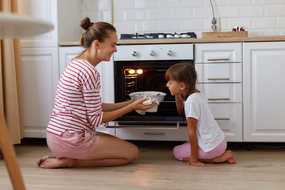 A mom shows her young daughter how to use the oven.