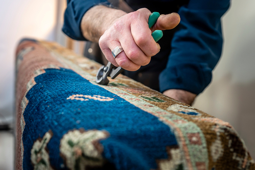 A rugmaker alters a blue rug using a special metal tool.